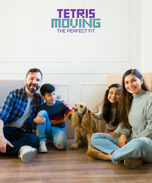 Family of 4 plus a dog sitting on a hardwood floor in front of moving boxes