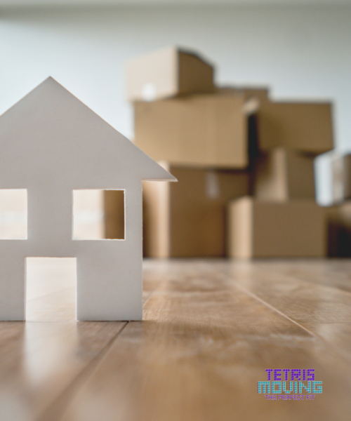 A small paper cutout of a house in the foreground with packing boxes in the background